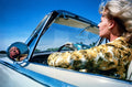 Blonde woman in floral jacket driving a vintage convertible car with Alfred Hitchcock's reflection in the side mirror under a clear blue sky