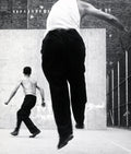High-contrast black and white photograph of two men playing handball on an outdoor urban court, featuring one man leaping mid-air.