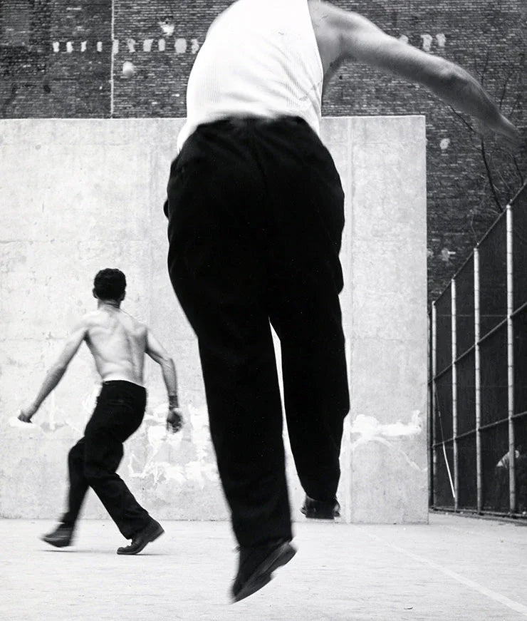 High-contrast black and white photograph of two men playing handball on an outdoor urban court, featuring one man leaping mid-air.