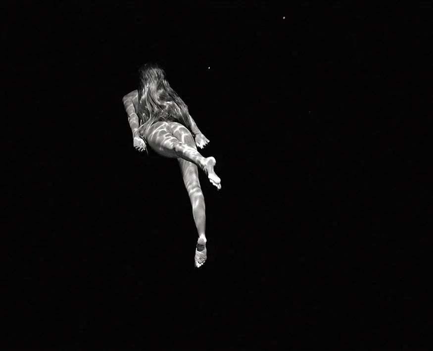 Black and white photo of a woman swimming underwater with light patterns on her body against a dark background