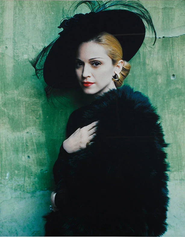 Madonna portrait in dramatic black fur, feathered hat, and red lipstick, posed against a distressed green wall.