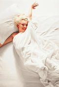 Candid color photograph of Marilyn Monroe smiling brightly while waking up, wrapped in white bedsheets with her arm raised.