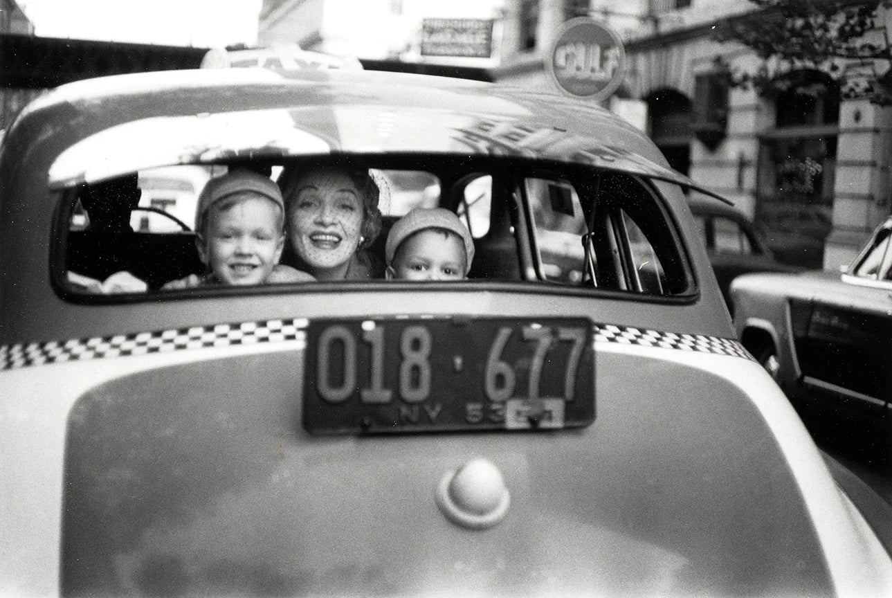 Black and white candid photo of Marlene Dietrich, smiling widely, looking out the rear window of a 1953 NYC taxi with her two young grandchildren.