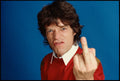 Iconic close-up portrait of Mick Jagger in a red sweater making an aggressive face and showing his middle finger against a bright blue studio backdrop.