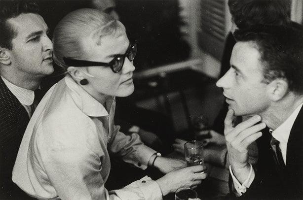 Candid black and white photograph showing Paul Newman, Joanne Woodward in cat-eye glasses, and Truman Capote chatting at a 1950s party.
