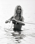 Brigitte Bardot in a striped bikini top pulling a rope while standing waist-deep in the ocean, black and white vintage photograph.