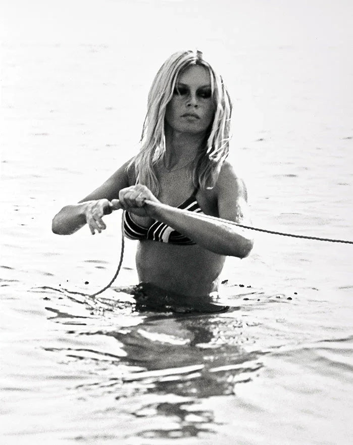 Brigitte Bardot in a striped bikini top pulling a rope while standing waist-deep in the ocean, black and white vintage photograph.