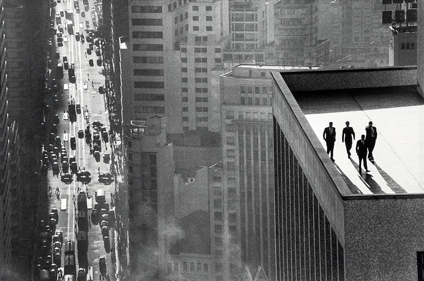 Dramatic black and white photograph showing four men in suits standing on a high skyscraper ledge overlooking a busy New York City street.