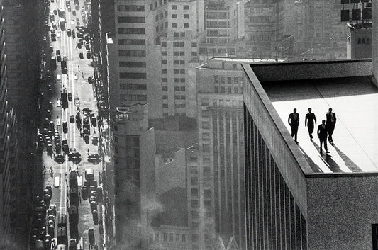 Dramatic black and white photograph showing four men in suits standing on a high skyscraper ledge overlooking a busy New York City street.