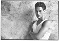 Black and white photo of Rob Lowe, looking intensely at the camera while leaning against a rough stucco wall in a white tank top.