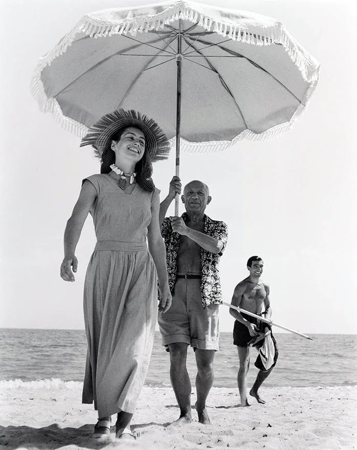 Black and white photo of three people on a beach with one holding a large umbrella over a smiling woman in a sunhat and long skirt