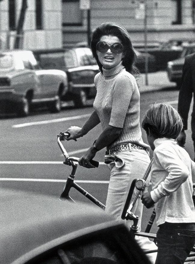 Black and white photo of stylish woman with sunglasses and boy holding bicycles on city street