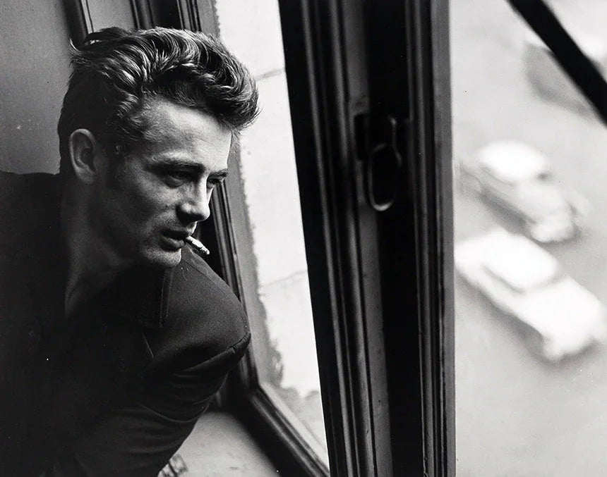 Black and white photo of James Dean, smoking a cigarette, leaning out of a city window looking down at the street traffic below.
