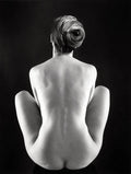 High-contrast black and white photograph of a female nude form seated from the back, knees drawn up, hair styled in a distinct swirl.