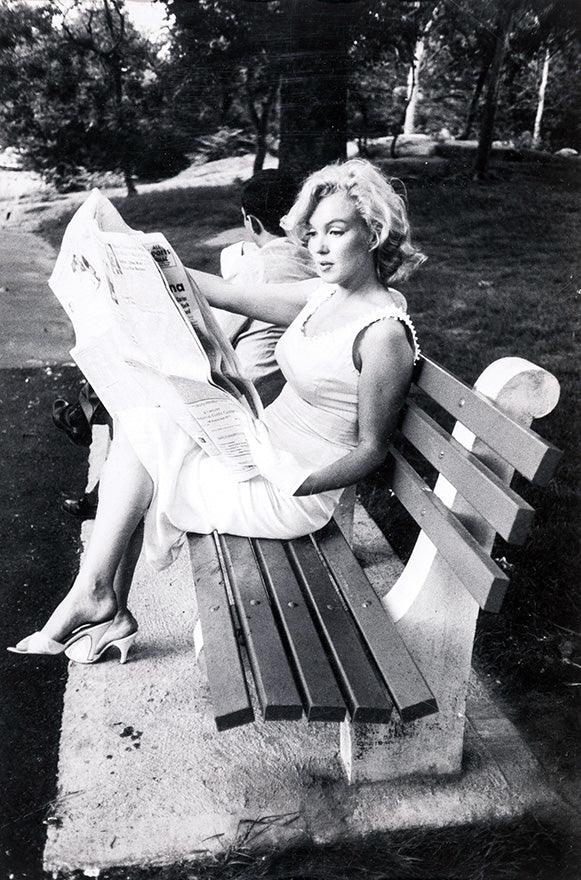 Black and white vintage photograph of Marilyn Monroe sitting on a wooden park bench reading a newspaper, wearing a white dress and heels.