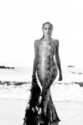 Black and white portrait of Sharon Stone wearing a form-fitting snakeskin gown while holding seaweed on a bright, sunny beach.
