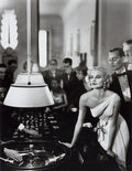 High contrast B&W photo of a glamorous blonde woman in a white gown leaning over a spinning roulette wheel in a busy 1950s luxury casino scene.