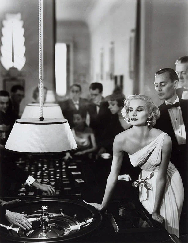 High contrast B&W photo of a glamorous blonde woman in a white gown leaning over a spinning roulette wheel in a busy 1950s luxury casino scene.