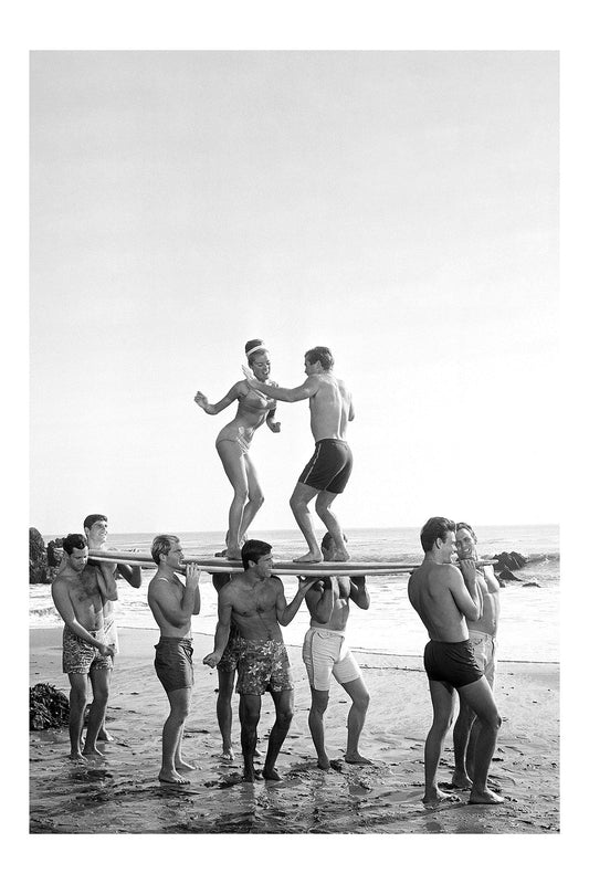 Black and white vintage photograph of eight young people on a sunny beach, with a man and woman balancing on a long surfboard carried horizontally on the shoulders of six men.