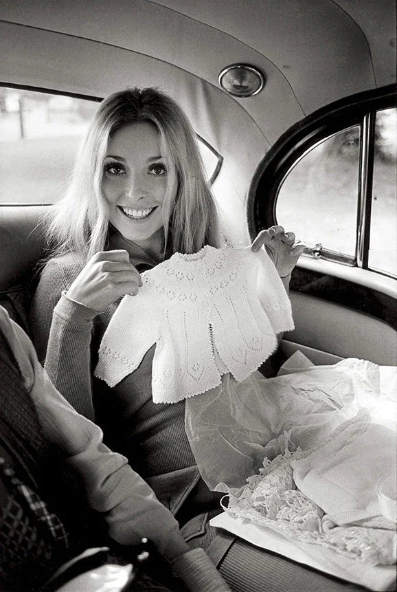 Black and white portrait of actress Sharon Tate smiling brightly while holding a delicate white knitted baby sweater inside a car.