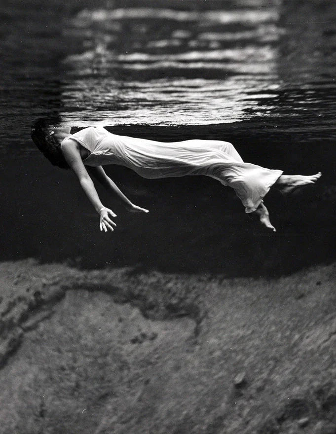 Black and white fine art photograph of a woman wearing a flowing white dress, peacefully floating just beneath the water's surface.