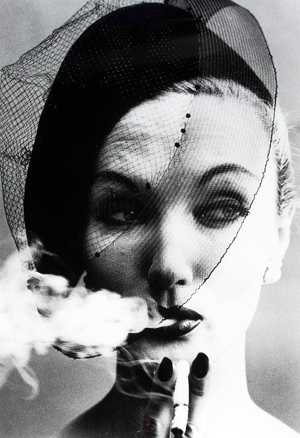 Black and white portrait of a woman wearing a netted veil hat, exhaling smoke with a cigarette in hand