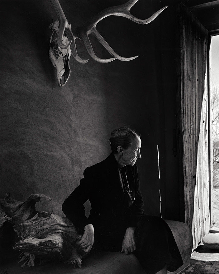 Black and white portrait of a woman sitting in a rustic room with antlers mounted on the wall and light from an open door
